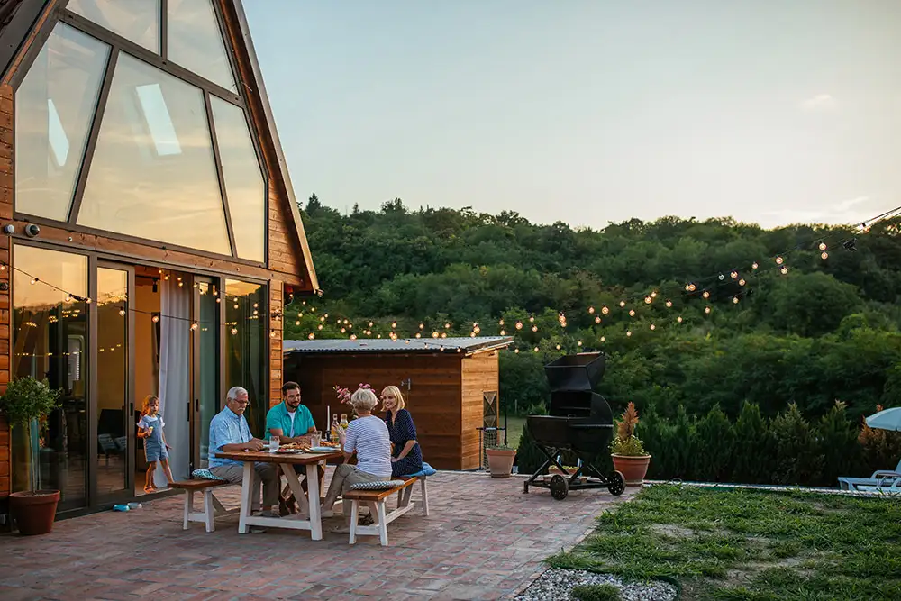 Family enjoying an outdoor dinner on a patio near a wooden house with large windows, surrounded by string lights and greenery.